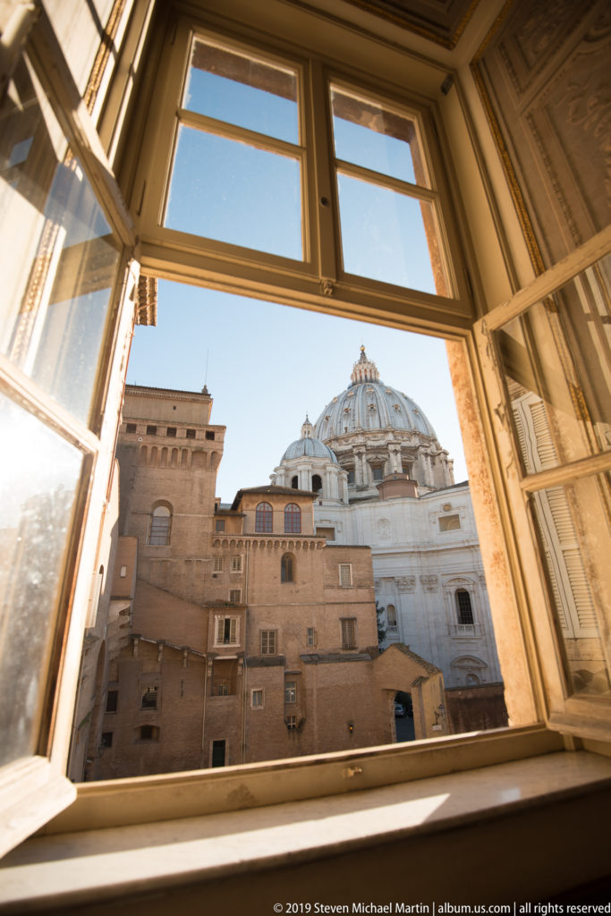 St. Peter's Basilica dome and surrounding Vatican buildings seen through an open wooden museum window under blue sky.