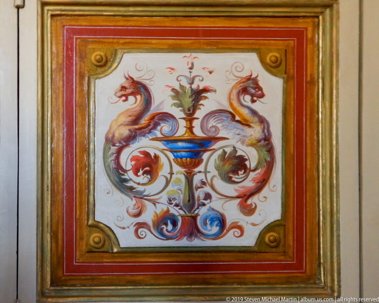 Painted decorative panel with a central vase and plant, paired dragons, colorful scrolling foliage, red border, and gilded frame.