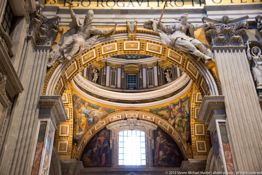 Ornate interior arch with gilded coffers, sacred mosaics, sculpted figures, Corinthian supports, and a bright window below.