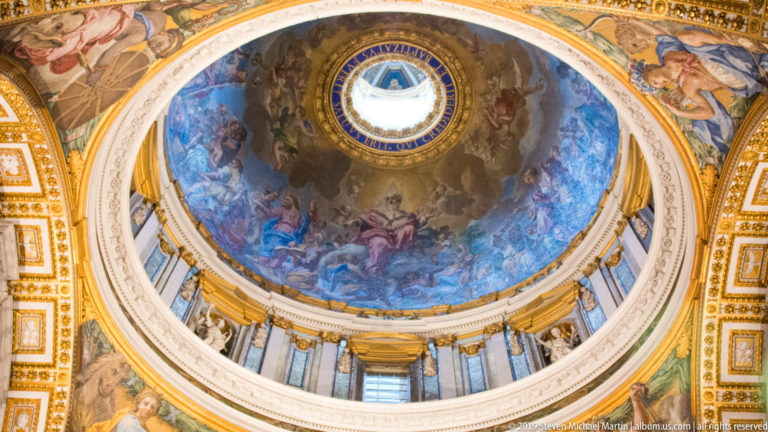 Upward view of a painted cupola with bright central oculus, cloud-filled figures, drum windows, gilded borders, and decorated pendentives.