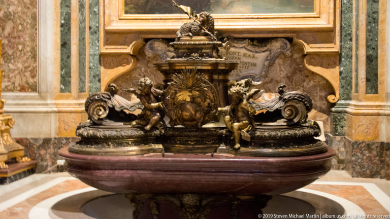 Ornate bronze basin with central heraldic shield, scrolling forms, winged putti, and Latin inscription on a reddish marble support.