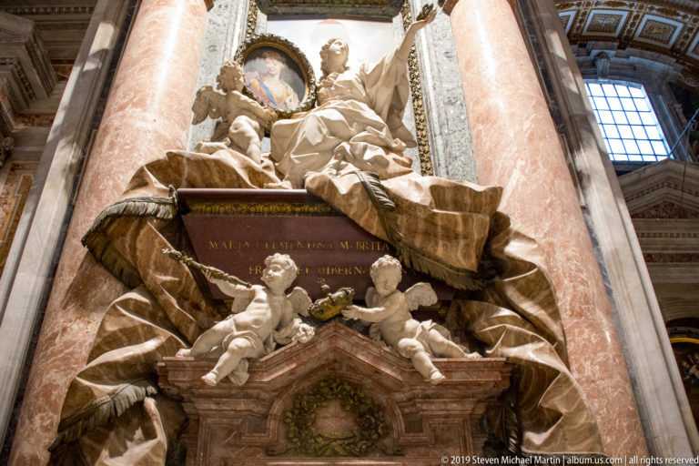 Low-angle view of a marble memorial with a seated female figure above two winged putti, inscription panel, and red marble columns.