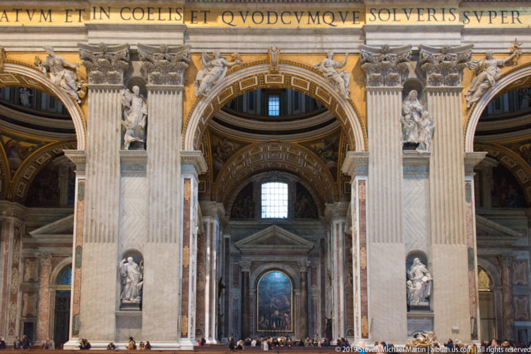 Centered interior bay with colossal fluted pilasters, saint statues in niches, decorated arches, gilding, and visitors below.