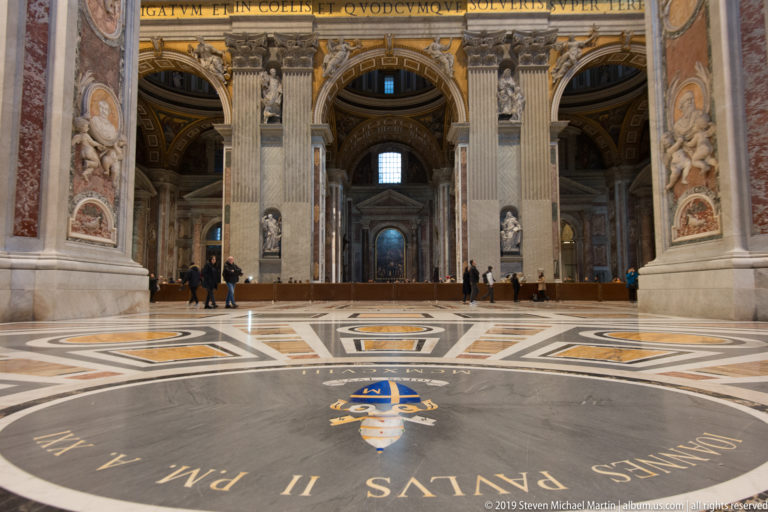 Wide interior view with giant pilasters, arches, visitors, and a large circular heraldic inscription in the marble pavement.