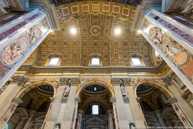 Steep upward view of giant pilasters and arches rising to a gilded coffered ceiling with windows and sculptural decoration.