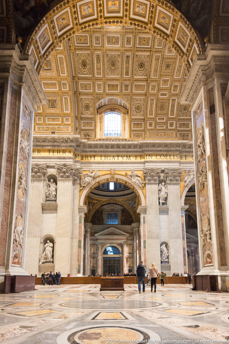 Wide interior view with gilded coffered ceiling, monumental piers and arches, patterned marble floor, niches, and scattered visitors.