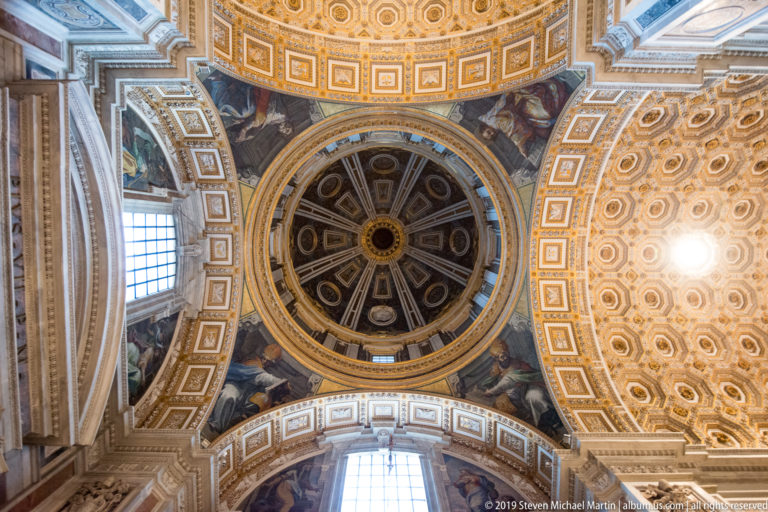 Upward view of a small ribbed cupola surrounded by painted pendentives, gilded coffers, windows, and ornate vault decoration.