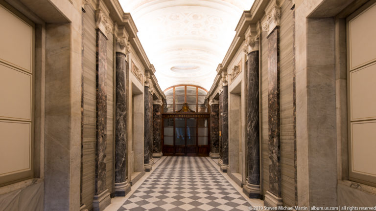 Long marble corridor with black-and-white diamond floor, repeated piers, pale vaulted ceiling, and dark doorway at the far end.