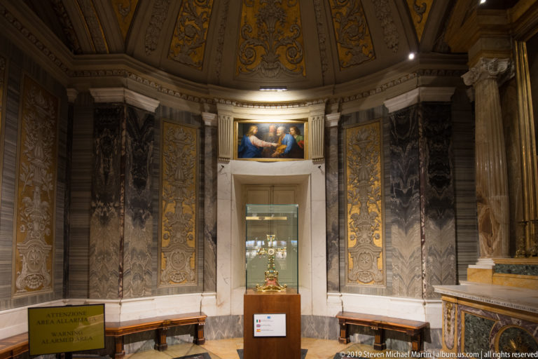 Small domed chapel with dark marble walls, gilded panels, benches, a central glass display case, and a sacred painting above.