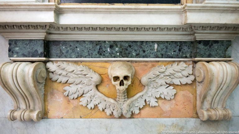 Pale carved skull with outstretched feathered wings centered on a pink marble panel between scroll-shaped supports.