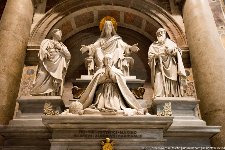 Marble monument with a kneeling pope below enthroned Christ, flanked by two standing saints in an arched niche.