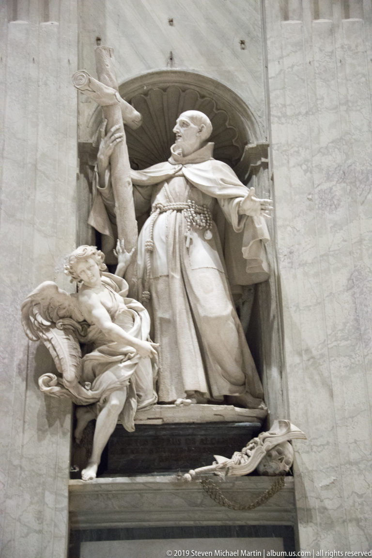 Large marble papal monument with a standing pope raising one hand, accompanied by smaller allegorical figures in a deep niche.