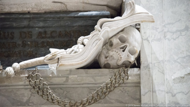 Marble skull emerging beneath carved drapery at a funerary monument, with chain and skeletal details nearby.