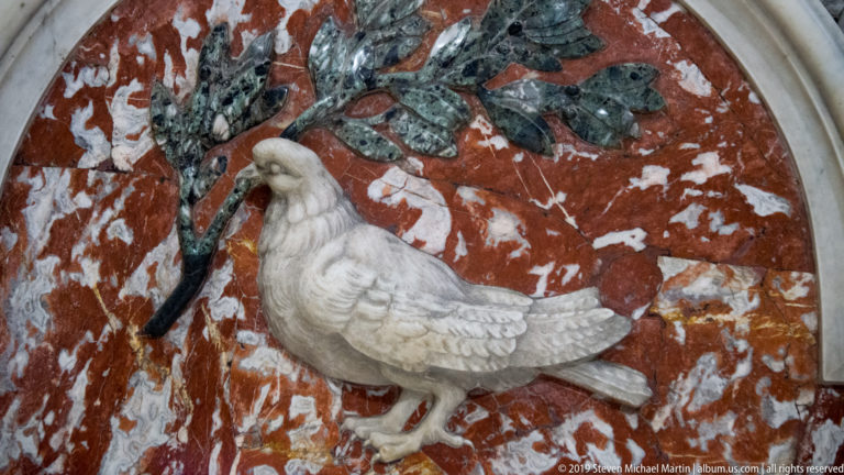 White marble dove holding a leafy branch in its beak against a red and green veined stone background.