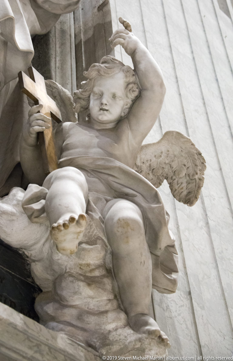 Winged marble putto seated on a monument, holding a small cross and lifting one arm overhead.