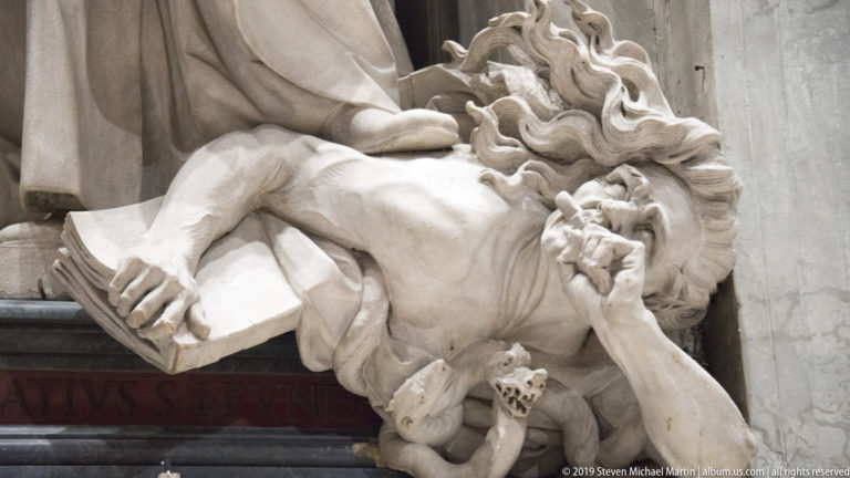 Reclining bearded marble figure holding an open book, with deeply carved hair and dramatic drapery.