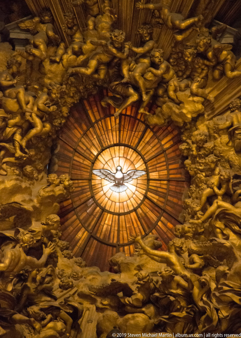 Close view of the Holy Spirit dove in a radiant amber window surrounded by dense gilded clouds and angelic figures.