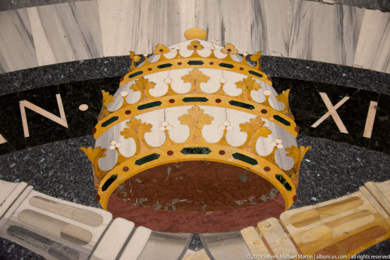 Polychrome marble floor inlay shaped as a papal tiara with gold, white, red, green, and black stone.