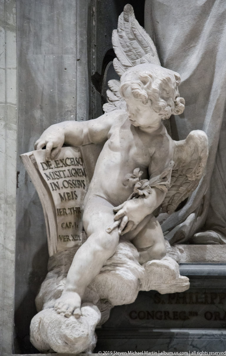 Winged marble putto reclining beside an open book carved with Latin text.