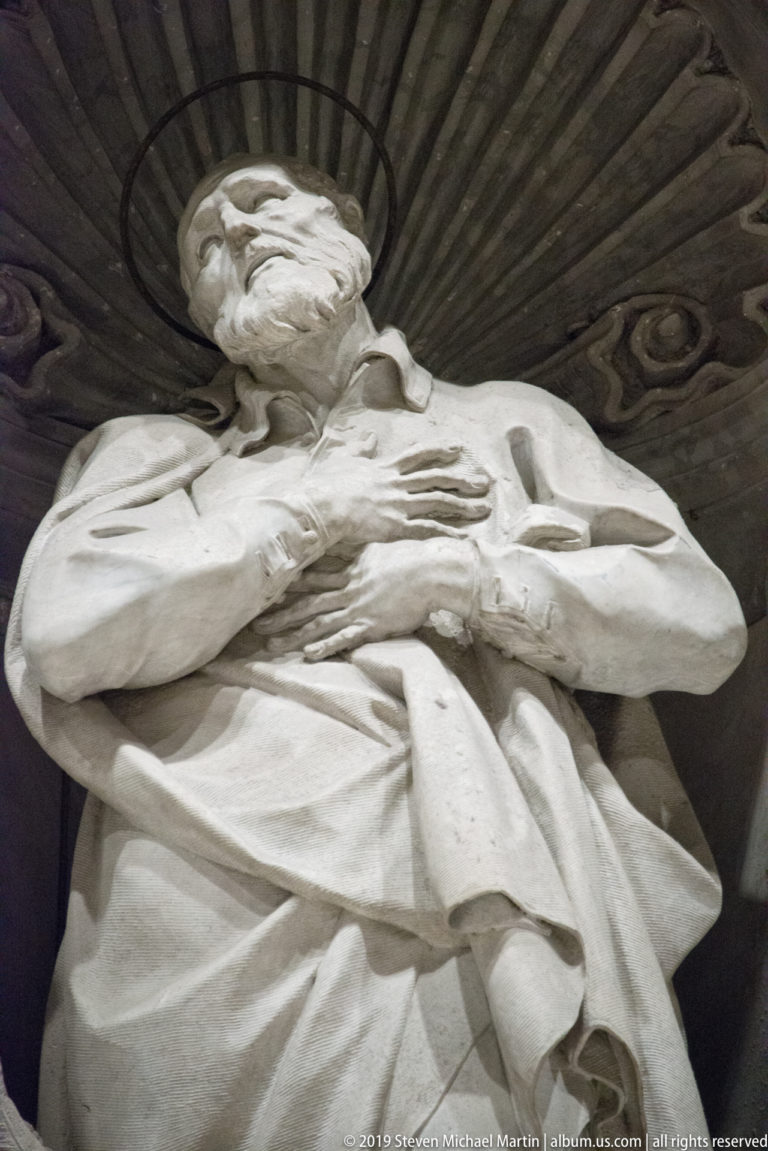 Low-angle close view of a bearded marble saint with crossed hands and sweeping robes beneath a shell-shaped canopy.