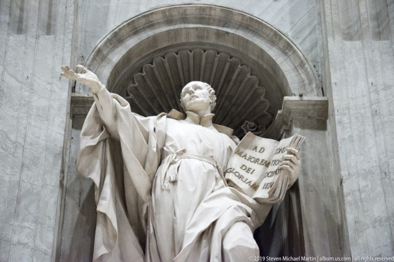 Monumental marble saint in a shell niche, extending one arm while holding an open inscribed book.