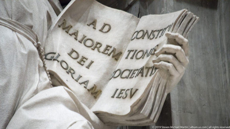 Marble hands hold an open sculpted book with large incised Latin words across its pages.