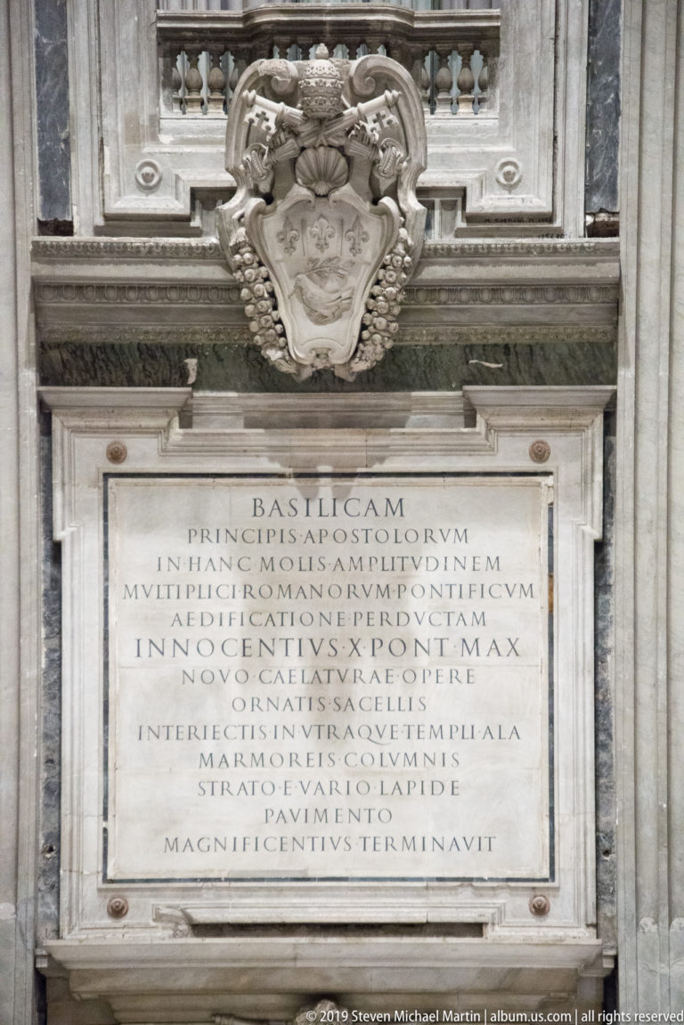 Latin inscription tablet in a marble frame beneath an ornate papal coat of arms.