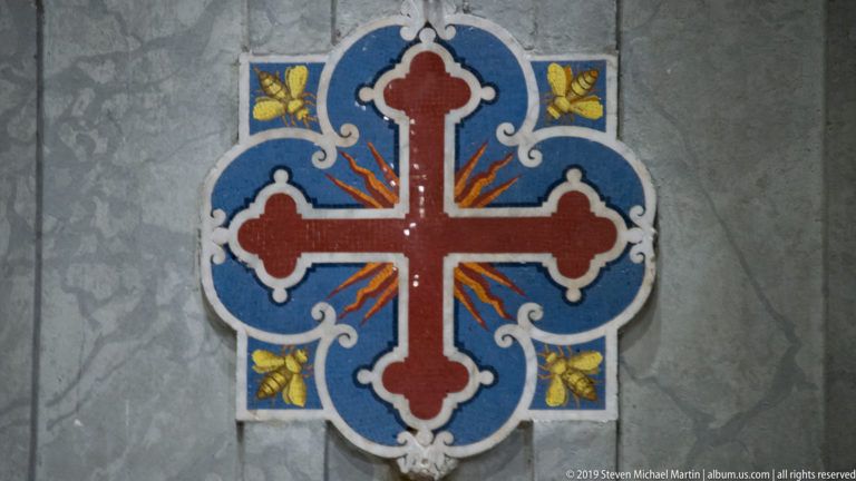 Red cross within a blue quatrefoil ornament with white scrollwork and gold foliage on gray marble.