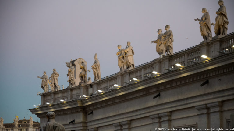 Angled dusk view of numerous stone saint statues standing along the illuminated balustrade above Bernini’s colonnade.