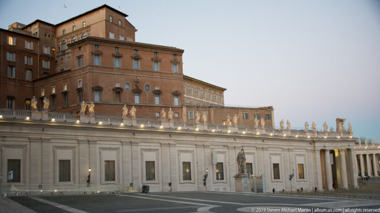 Closer evening view of the Apostolic Palace above the colonnade wall and rooftop saints, with patterned paving across Saint Peter’s Square.