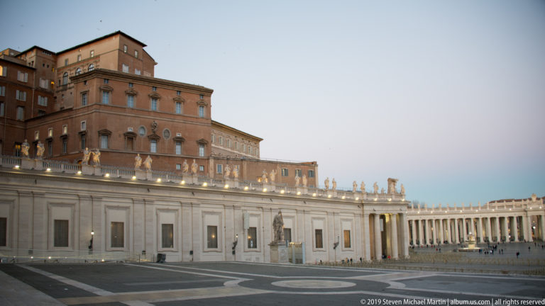 Apostolic Palace rising behind the pale colonnade wall and rooftop statues of Saint Peter’s Square, with curved paving patterns in the foreground.