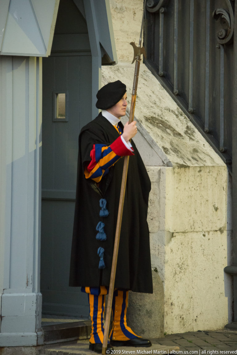 Swiss Guard in profile holding a tall halberd, wearing a dark cloak over blue, red, and yellow striped uniform beside a gateway.