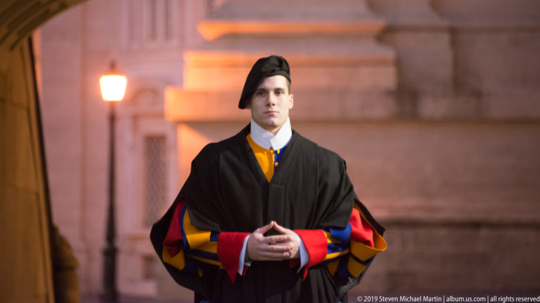 Portrait of a Pontifical Swiss Guard wearing a dark cloak over red, blue, and yellow ceremonial uniform, hands clasped before him.