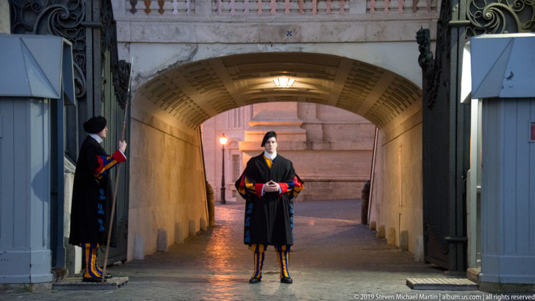Two Pontifical Swiss Guards in striped uniforms and dark cloaks stand at a stone gateway with a warmly lit arched passage behind them.