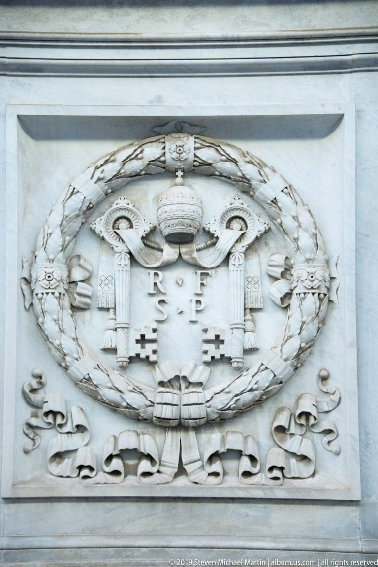 White marble relief of papal heraldry with tiara, crossed keys, shield, leafy wreath, and ribbon within a rectangular frame.