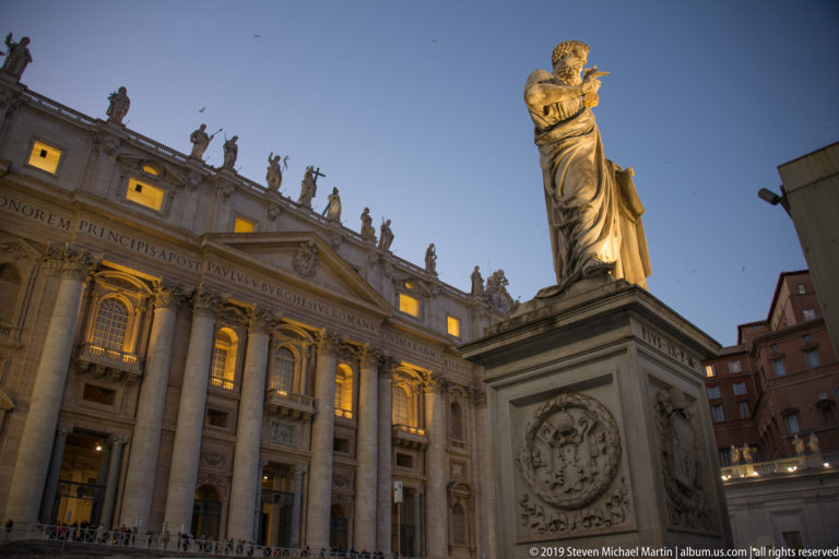 Colossal marble statue of Saint Paul on its pedestal before the illuminated façade of Saint Peter’s Basilica.