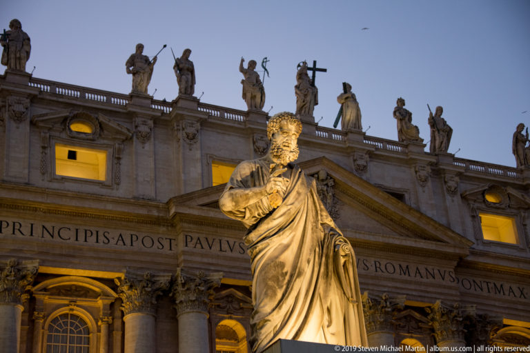 Illuminated stone statue of Saint Peter holding keys before the façade, inscription, and rooftop statues of Saint Peter’s Basilica.
