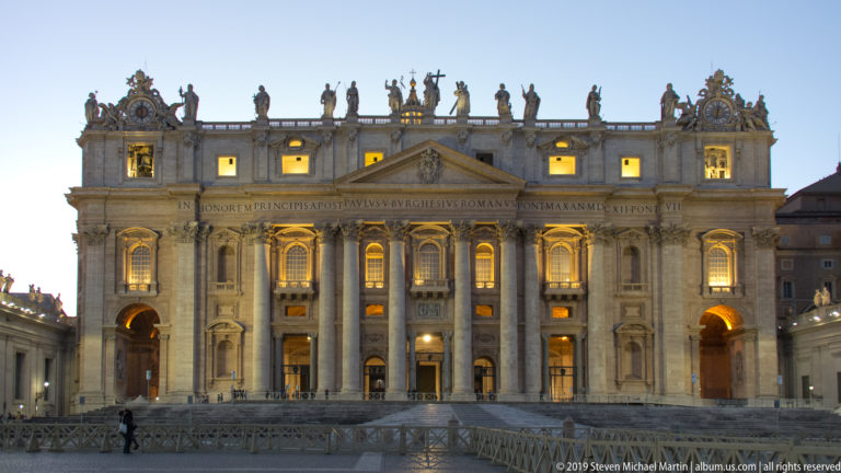 Symmetrical evening view of Saint Peter’s façade with central pediment, balcony, giant columns, glowing windows, inscription, and rooftop statues.