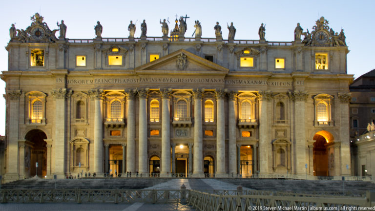 Closer frontal view of Saint Peter’s illuminated façade with colossal columns, portals, attic inscription, rooftop statues, and glowing windows.