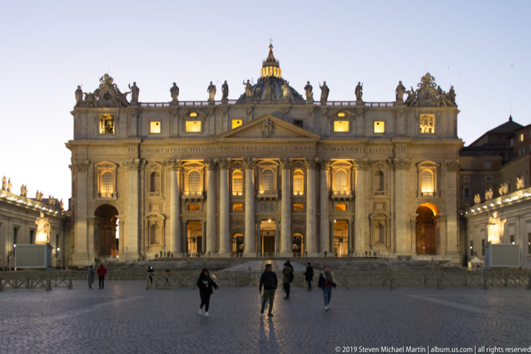 Centered blue-hour view of Saint Peter’s Basilica with illuminated façade, dome, rooftop statues, inscription, and visitors crossing the square.
