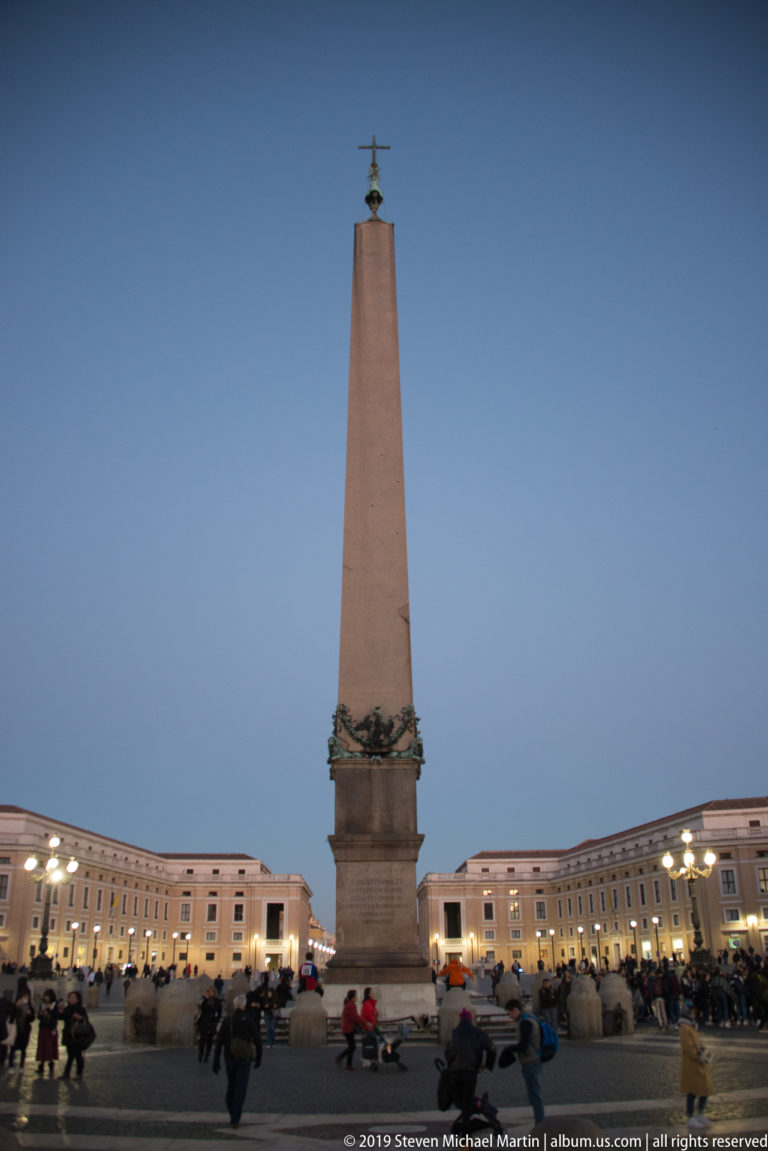 Centered view of the Vatican Obelisk on its stone pedestal, crowned by a cross against a blue-hour sky with visitors below.