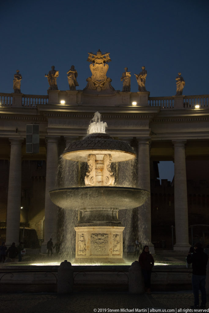 Illuminated tiered fountain with water cascading into a broad basin, backed by the colonnade and rooftop statues of Saint Peter’s Square.