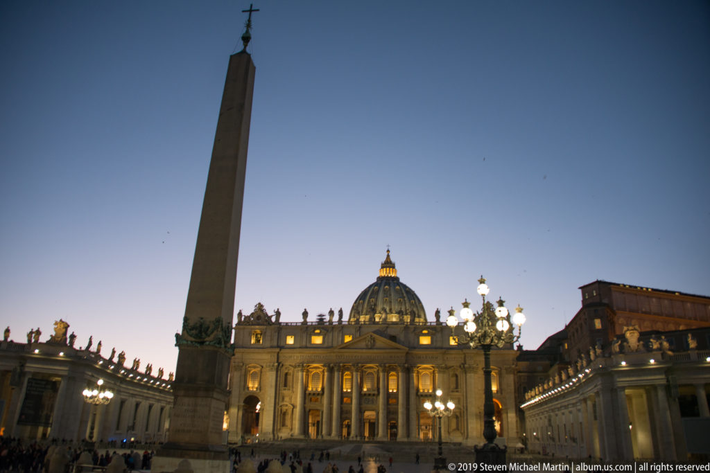 Vatican Obelisk rising before the illuminated façade and dome of Saint Peter’s Basilica, with colonnades and glowing lamps at dusk.