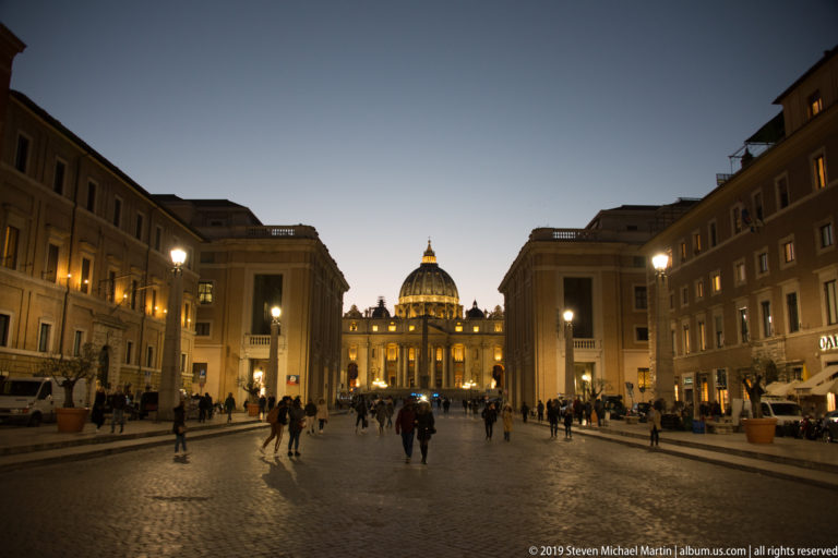 Broad dusk view down Via della Conciliazione toward the illuminated façade and dome of Saint Peter’s Basilica, with pedestrians and glowing streetlights.