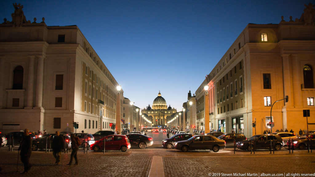 Dusk street view along Via della Conciliazione with cars, pedestrians, lit buildings, and Saint Peter’s illuminated dome centered in the distance.