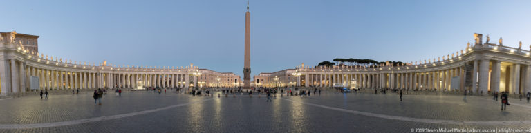 Wide panoramic view across Saint Peter’s Square with both curving colonnades, the central Vatican Obelisk, evening lights, and visitors.