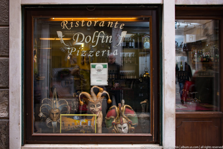 Restaurant window labeled Ristorante Dolfin Pizzeria with colorful Venetian carnival masks displayed behind the glass.
