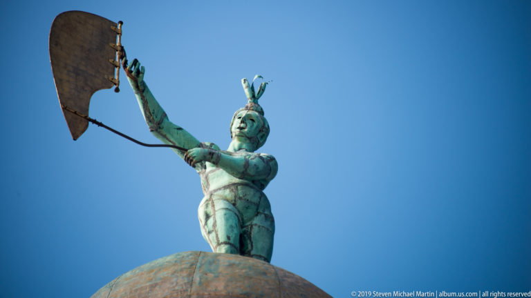 Bronze Fortuna figure holding a broad sail above a rounded globe against a clear blue sky.