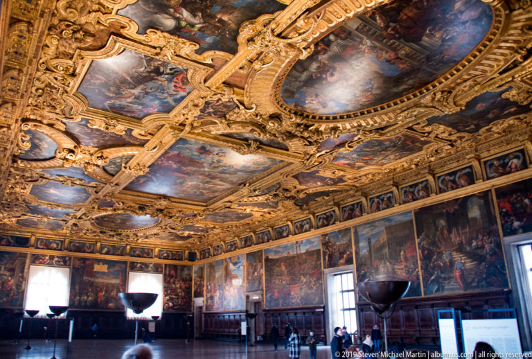 Large palace hall with gilded compartmented ceiling, extensive wall paintings, tall windows, and visitors below.