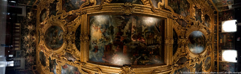 Corner of an ornate palace room with gilded carved frames surrounding large ceiling and wall paintings.
